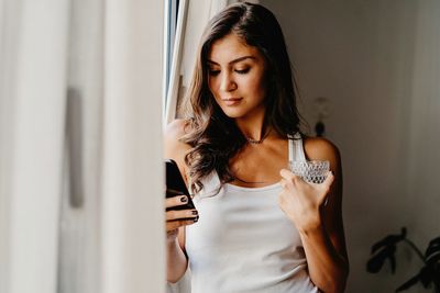 Positive millennial female in sleepwear holding glass of fresh water and reading news in social networks on smartphone while standing near window in bedroom in morning