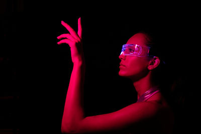 Side view of serious focused young female touching invisible object while experiencing virtual reality in modern transparent vr glasses in dark studio with bright pink neon illumination