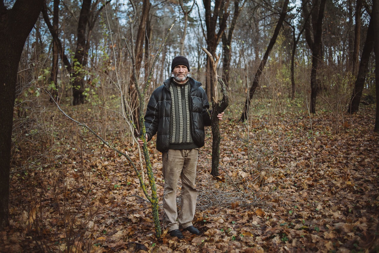 tree, forest, lifestyles, leisure activity, casual clothing, young adult, person, tree trunk, woodland, front view, standing, nature, tranquility, looking at camera, portrait, full length, leaf, outdoors