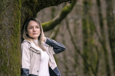 Woman in a light-colored coat leaning against a mossy tree