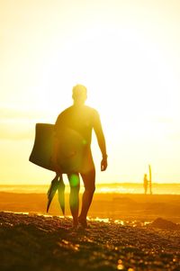 Full length of silhouette man carrying surfboard on shore at beach against sky during sunset