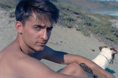 Portrait of shirtless young man with dog sitting on sand at beach