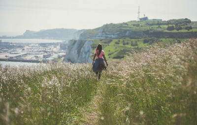 Rear view of woman walking on field by sea against sky