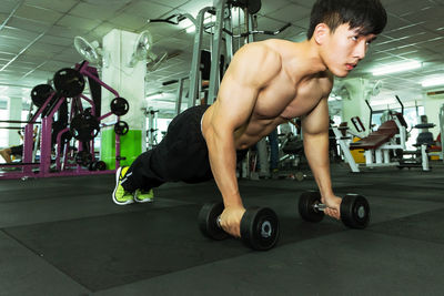 Full length of shirtless man lifting dumbbells at gym