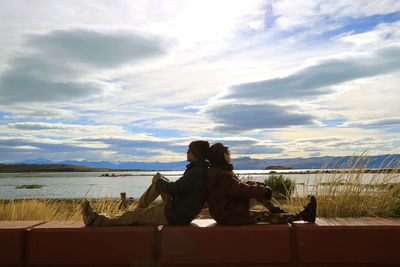 Side view of couple sitting on retaining wall by lake against cloudy sky