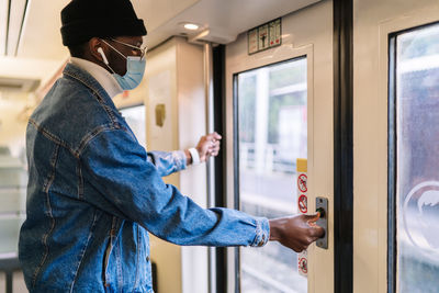 Side view of african american male passenger in mask standing at doors and pressing button before leaving train while traveling during coronavirus epidemic