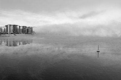 Lake by buildings against sky during foggy weather