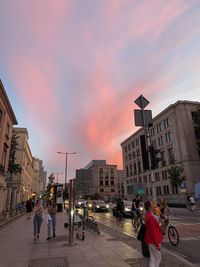 People walking on city street against sky at sunset