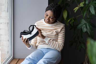 Young excited black woman sitting on windowsill looking curiously at vr headset in hands