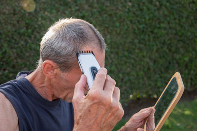 A gray-haired middle-aged man shaves his hair with a clipper in a garden