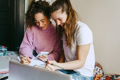 Happy young female friends using smart phone while homework