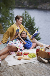 Female couple having picnic, reading book, and using phone