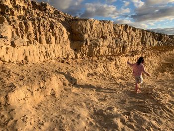 Rear view of girl playing at beach by rock formations against cloudy sky