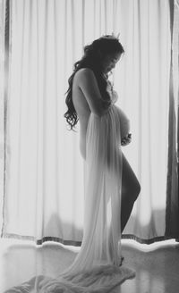 Side view of naked pregnant woman standing against curtain at home