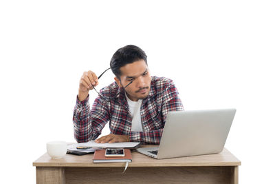 Young businessman holding eyeglasses while using laptop at desk against white background