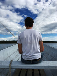 Rear view of man with fishing rod sitting on boat in lake against cloudy sky