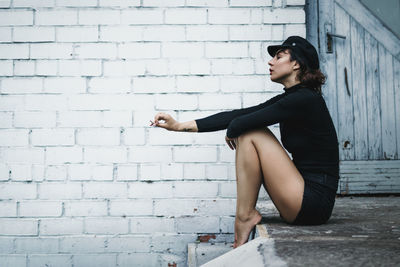 Side view of young woman sitting against brick wall