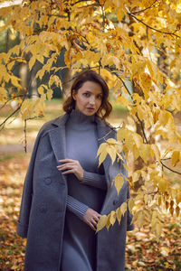 Brunette woman in a gray coat and boots is standing by a tree in autumn among the trees