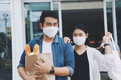 Portrait of smiling couple wearing flu mask holding shopping bag standing outdoors