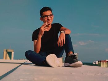 Smiling young man smoking cigarette while sitting on building terrace against blue sky