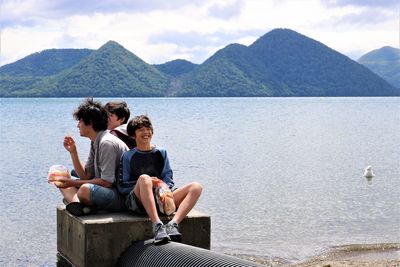 Rear view of friends sitting by lake against mountains