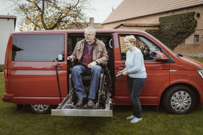 Smiling senior woman holding remote control while helping man with disability in motorized wheelchair to disembark from