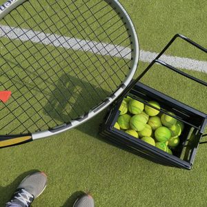 Low section of man standing with tennis racket by balls in container at court