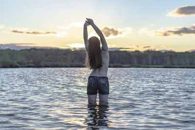 Rear view of woman with arms raised standing in lake against sky