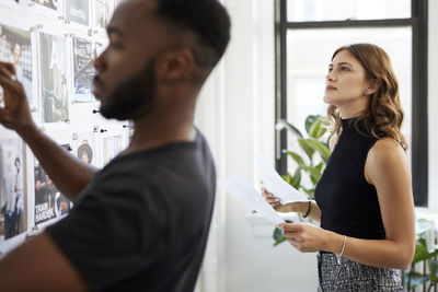 Businesswoman looking at photograph printouts on bulletin board while male colleague working in foreground