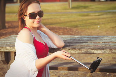 Side view of sensuous young woman holding monopod while taking selfie through smart phone on picnic table in park