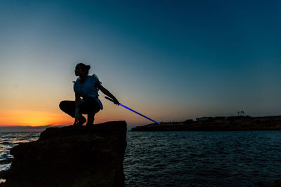 Silhouette man on rock by sea against sky during sunset