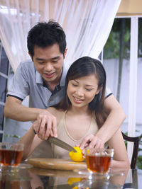 Man assisting woman in cutting fruit while having tea on glass table at home