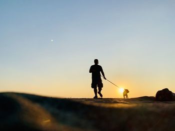 Silhouette man with dog walking on land against sky during sunset