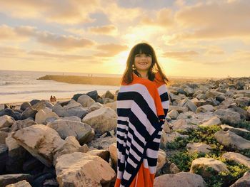 Smiling girl wrapped in towel while standing on rocks at beach against sky during sunset