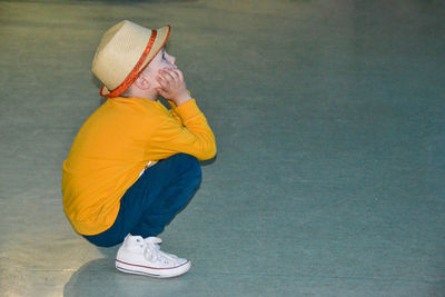 A boy wearing a yellow shirt and a hat is kneeling on a gravel road.