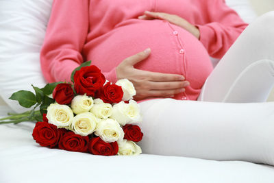 Midsection of pregnant woman sitting with rose bouquet on bed at home