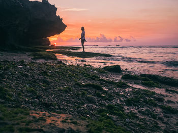 Man standing on rock by sea against sky during sunset