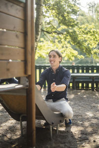 Smiling man waiting for baby boy while sitting on slide at playground in park
