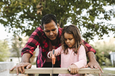 Girl cutting wooden plank by father during summer