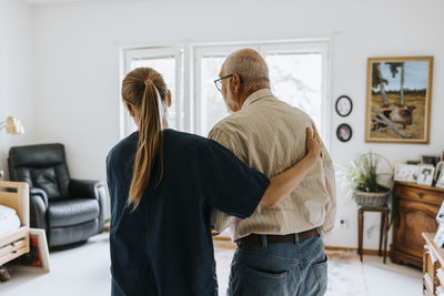 Rear view of female nurse supporting senior male while walking at elderly care home