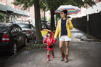 Happy mother and daughter holding umbrellas while walking on sidewalk