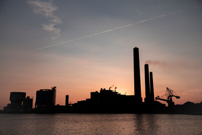 Silhouette smoke stacks by river in city during sunset
