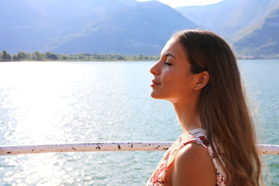 Side view of young woman with eyes closed by lake against mountains during sunny day