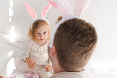 Man and girl, kid wearing bunny ears playing with pink eggs on bed by white wall at home.