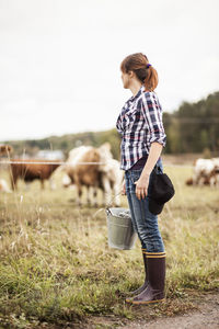 Female farmer with bucket standing on field with animals grazing in background