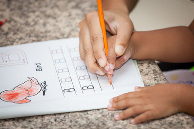 Cropped hand of parent assisting child writing on book at table
