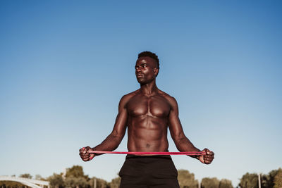 Male athlete looking away while stretching resistance band standing against clear sky during sunset