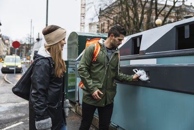 Heterosexual couple throwing plastic in recycling bin while standing at city