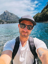 A man wearing sunglasses and a hat is taking a selfie in front of a body of water. 