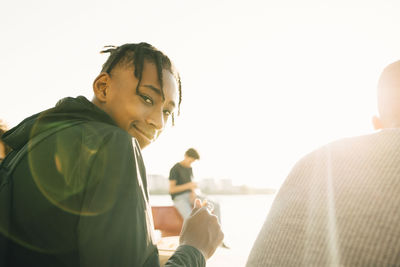 Portrait of smiling teenage boy holding meal while sitting by friend on promenade during sunny day